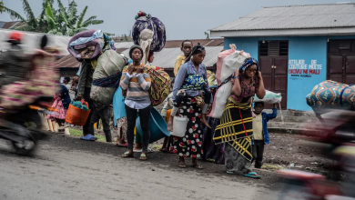 People carrying belongings on their heads walk along a road, with blurred motorcycles passing by.