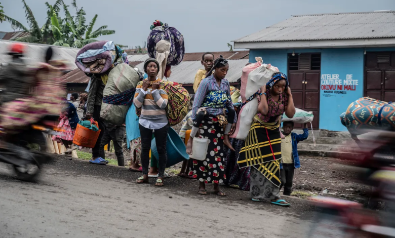 People carrying belongings on their heads walk along a road, with blurred motorcycles passing by.