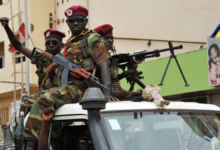 Soldiers in camouflage uniforms and berets ride on a vehicle armed with mounted machine guns in an urban setting.