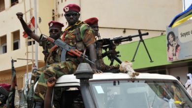 Soldiers in camouflage uniforms and berets ride on a vehicle armed with mounted machine guns in an urban setting.