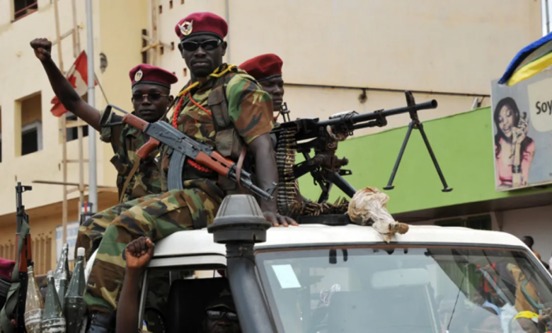 Soldiers in camouflage uniforms and berets ride on a vehicle armed with mounted machine guns in an urban setting.