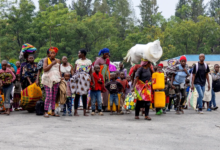 A group of people walking, carrying bags and containers. Trees and buildings are in the background.