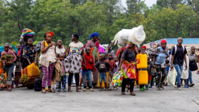 A group of people walking, carrying bags and containers. Trees and buildings are in the background.