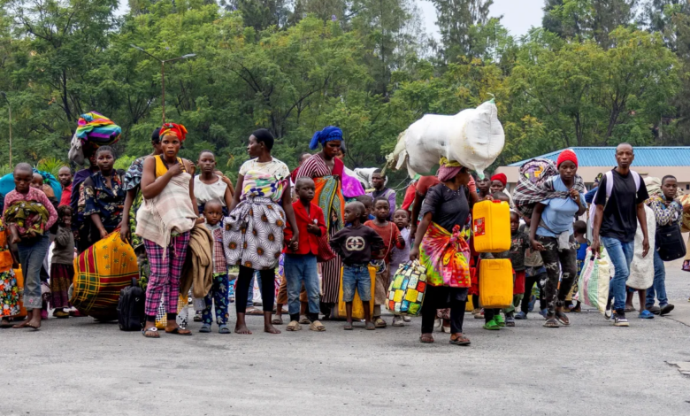 A group of people walking, carrying bags and containers. Trees and buildings are in the background.