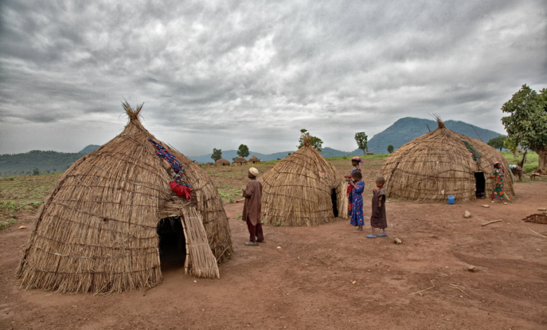 Thatched huts under cloudy skies, with people standing outside. Mountains and trees in the background.