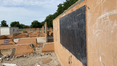 Outdoor classroom ruins with a chalkboard, surrounded by debris and greenery in the background.