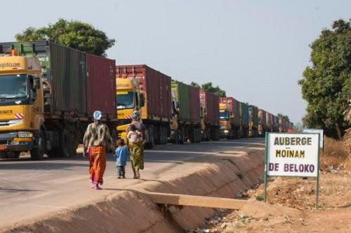 Long line of trucks on a road near trees, with people walking and a sign reading "Auberge Moinam de Beloko" beside them.