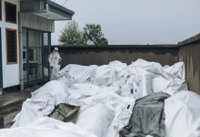 Person in protective gear spraying disinfectant on numerous white body bags outside a building.