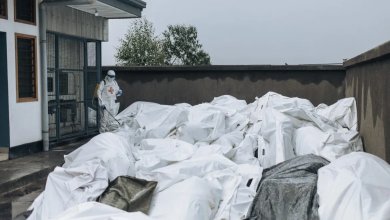 Person in protective gear spraying disinfectant on numerous white body bags outside a building.