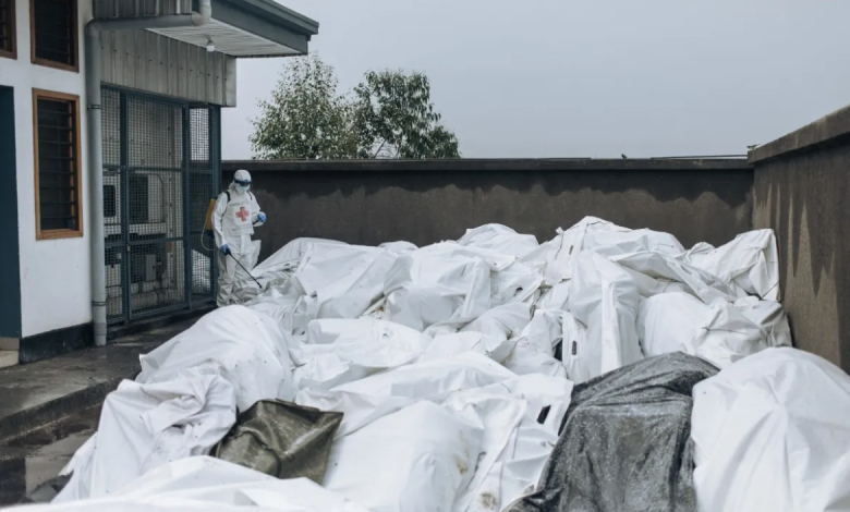 Person in protective gear spraying disinfectant on numerous white body bags outside a building.