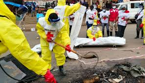 Emergency workers in yellow protective suits dig through debris while others in white vests observe, with a stretcher in the background.