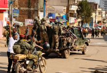 Soldiers in camouflage uniforms on a truck, with armed personnel on a motorcycle, in a busy urban area with buildings and people nearby.