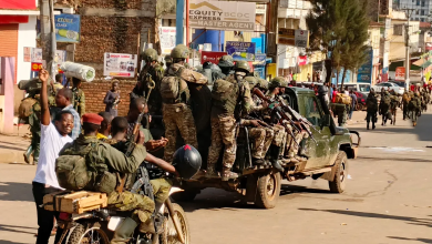 Soldiers in camouflage uniforms on a truck, with armed personnel on a motorcycle, in a busy urban area with buildings and people nearby.