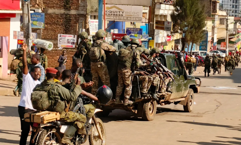 Soldiers in camouflage uniforms on a truck, with armed personnel on a motorcycle, in a busy urban area with buildings and people nearby.