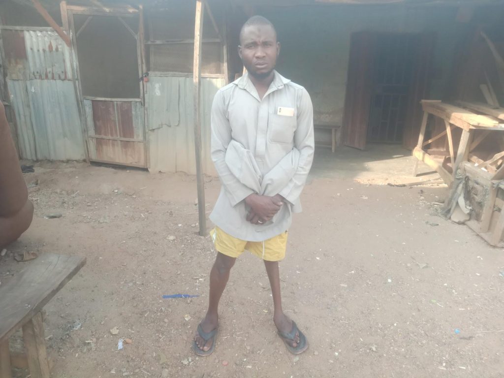 Man in a light shirt and yellow shorts stands outside a rustic building on a dirt ground.