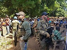 Armed security personnel stand guard as a crowd gathers in a shaded outdoor area.