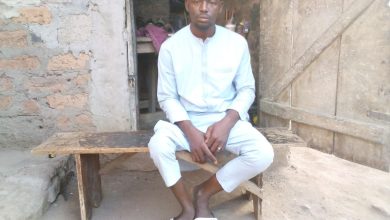 A man in a light outfit sits on a bench outside a rustic building, looking at the camera.