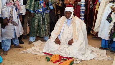 A man in traditional robes kneels on the ground, surrounded by others in colorful attire.