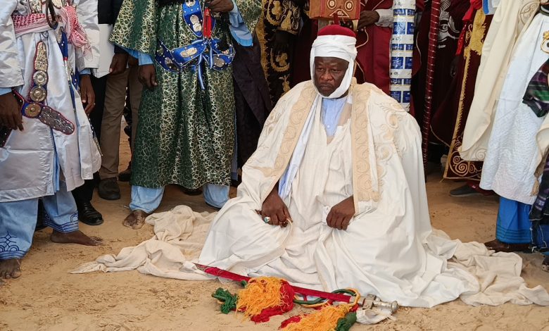 A man in traditional robes kneels on the ground, surrounded by others in colorful attire.