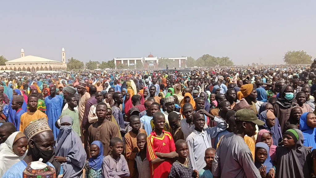 A large crowd gathers outdoors on a clear day, with people wearing colorful clothing and a mosque visible in the background.