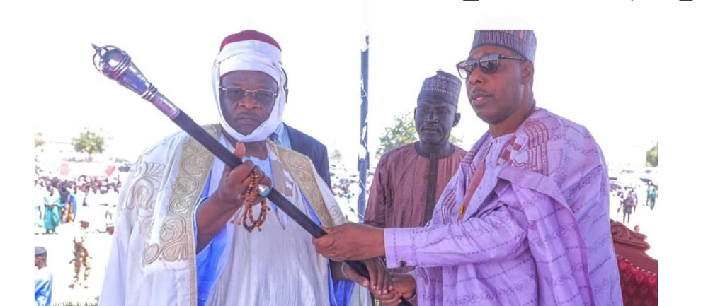 Two men in traditional African attire hold a staff, with a crowd in the background.