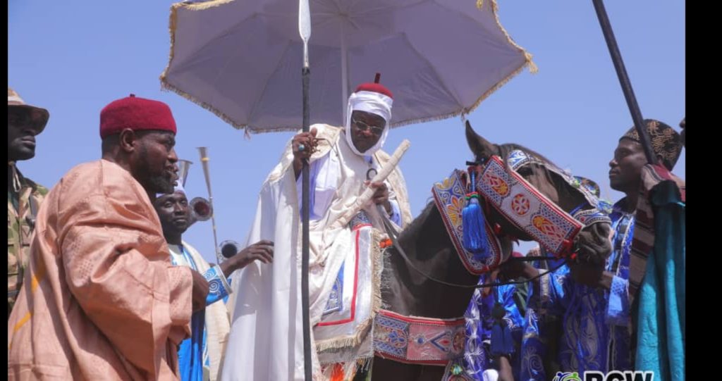 A person in traditional attire sits on a decorated horse, shaded by a large white umbrella, surrounded by others in festive clothing.