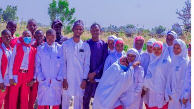 Group of students and teachers pose outdoors, some in red and others in white attire, smiling with greenery in the background.