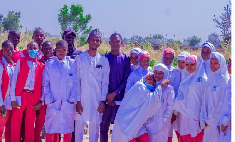 Group of students and teachers pose outdoors, some in red and others in white attire, smiling with greenery in the background.