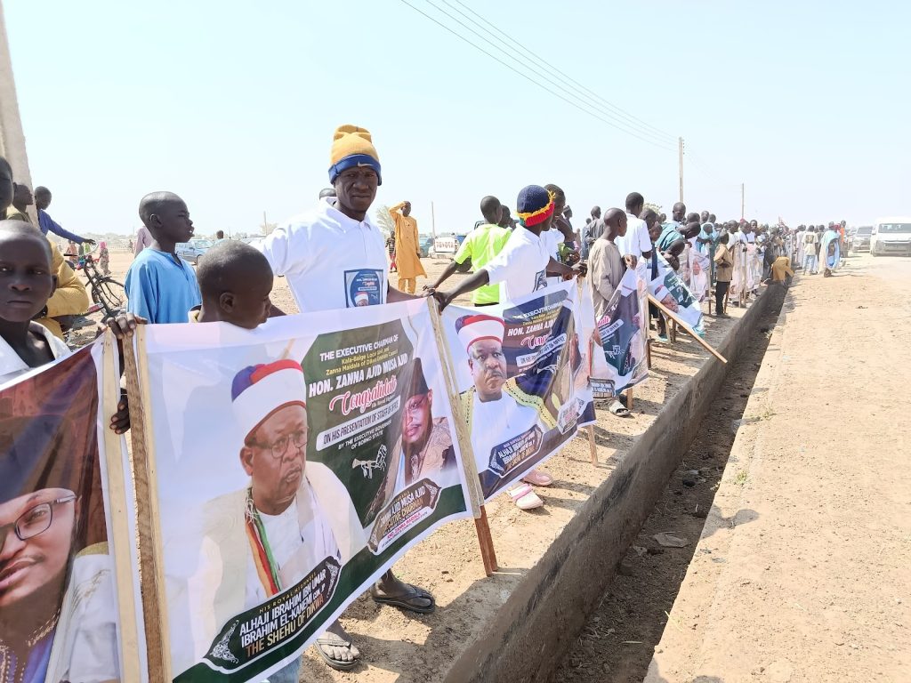 A group of people, some holding banners with images and text, stand along a road during a daytime event.