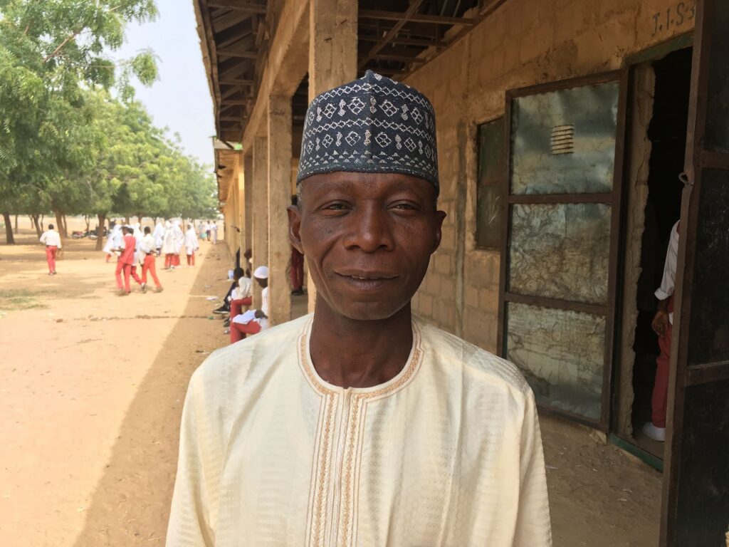 A man in a traditional cap and tunic stands outside a building, with students in uniforms in the background on a sunny day.