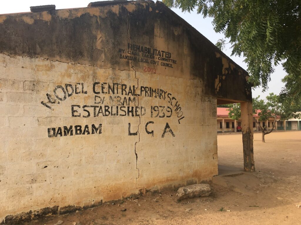 Exterior of Model Central Primary School, Dambam, with tree and surrounding buildings visible.