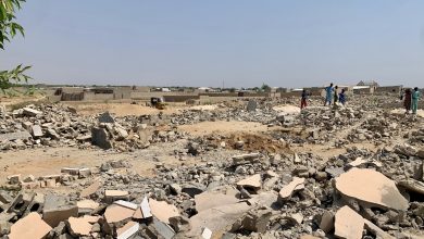 Rubble and debris in an open area with a few people and a small vehicle in the distance, under a clear sky.