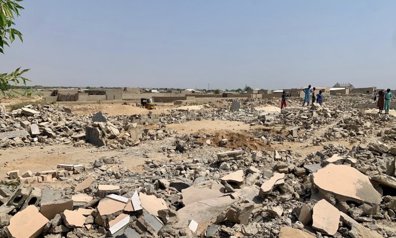 Rubble and debris in an open area with a few people and a small vehicle in the distance, under a clear sky.