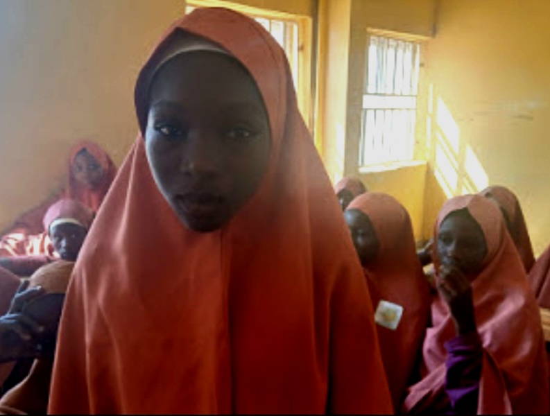 Girls wearing orange hijabs sit in a sunlit classroom.