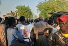 A group of people gathered around a Nigerian Red Cross worker outdoors, with trees and a vehicle in the background.