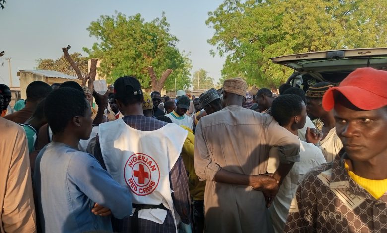 A group of people gathered around a Nigerian Red Cross worker outdoors, with trees and a vehicle in the background.