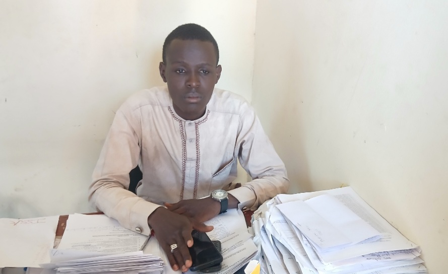 A person in a light shirt sits at a desk with stacks of papers, looking at the camera.