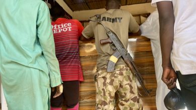 Four young boys stand at a wooden counter. One in army attire has a rifle slung over his back.