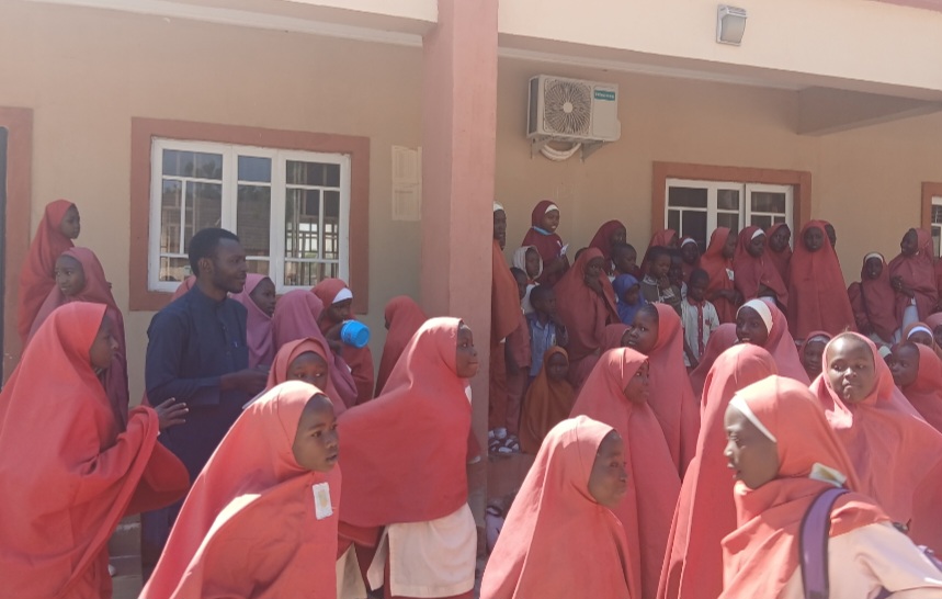 Group of children and a teacher, mostly in red uniforms, gathered outside a building with windows and an air conditioner.