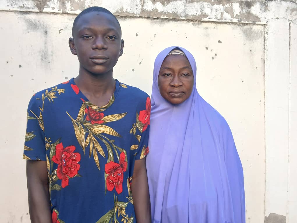 A young man in a floral shirt stands next to a woman in a purple hijab against a plain wall.