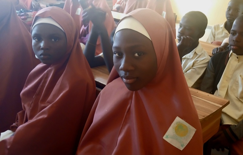 Students in maroon hijabs sit in a classroom, showing attentive expressions.