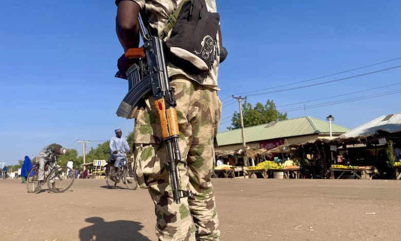 Person in camouflage with a rifle stands in a market street, with cyclists and market stalls in the background under a clear blue sky.