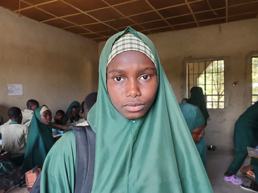 Young girl in a green hijab stands in a classroom with students in similar attire, sitting and working in the background.