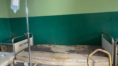 Worn hospital bed with damaged mattress in a green-walled room, next to an IV stand.