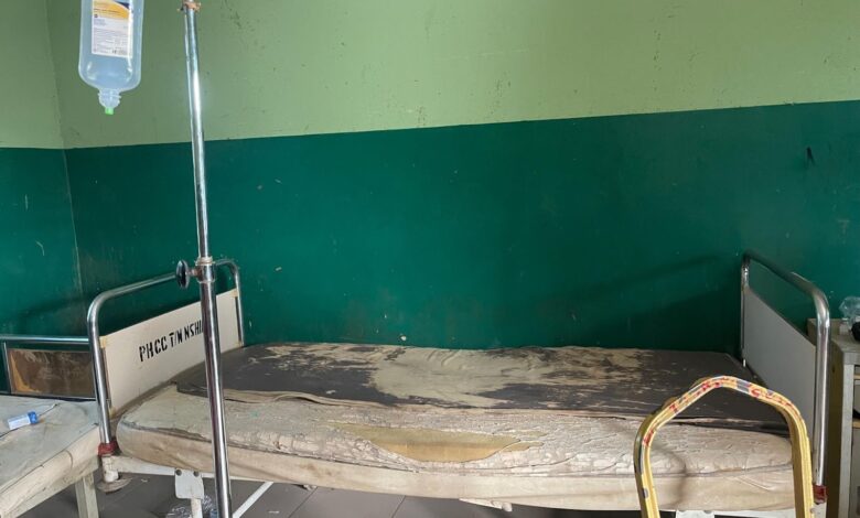 Worn hospital bed with damaged mattress in a green-walled room, next to an IV stand.