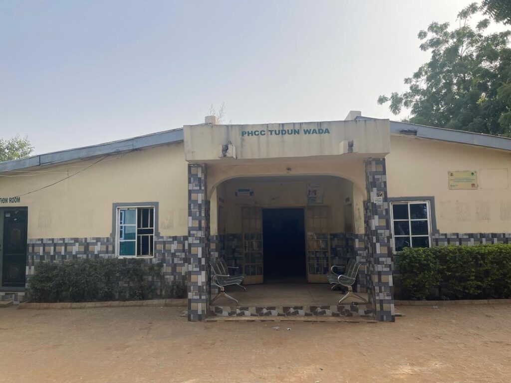 Exterior of a building labeled "PHCC Tudun Wada" with a tiled entrance and two metal benches outside.