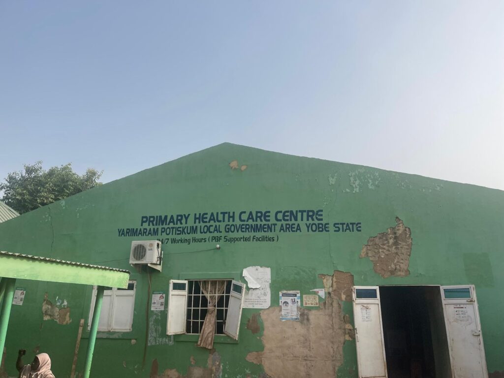 Green building with signs for "Primary Health Care Centre" in Yobe State. The exterior shows signs of wear, with open doors and windows.