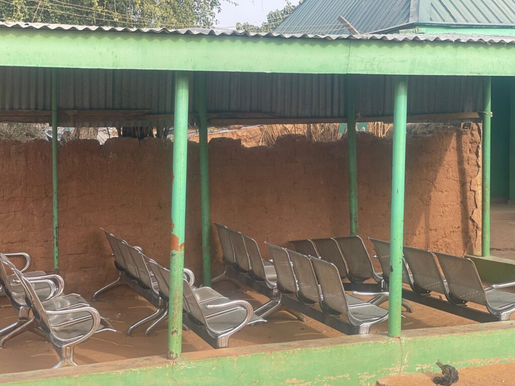 Outdoor waiting area with empty metal seats under a green roof, next to a mud wall.