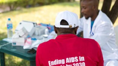 Person in red shirt with "Ending AIDS in Nigeria by 2030" sits with doctor outdoors, discussing at a table with medical supplies.