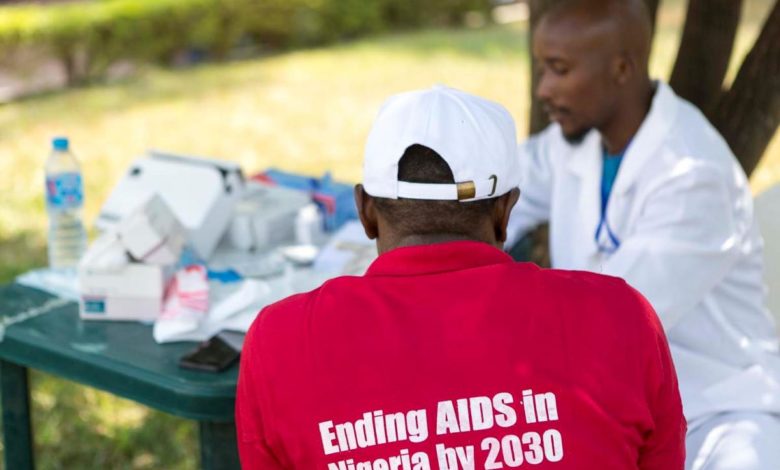 Person in red shirt with "Ending AIDS in Nigeria by 2030" sits with doctor outdoors, discussing at a table with medical supplies.